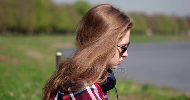 Tizenéves lány alkalmi napszemüveg hallgatta a zenét, ül, és nyugodt, zöld füvön, a folyó közelében — Stock videók