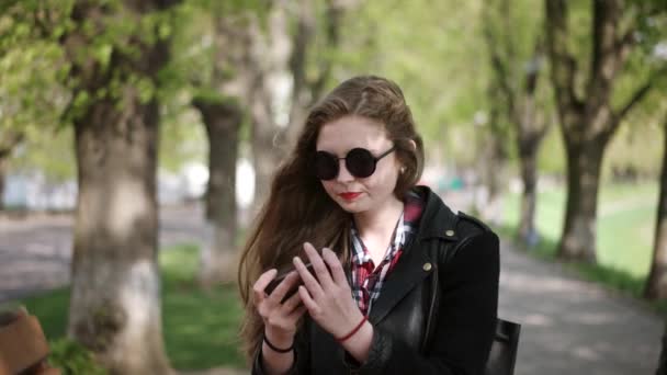 Adolescente urbana Casual Hipster Girl en gafas de sol usando un teléfono inteligente mientras está de pie en la pasarela de la ciudad — Vídeos de Stock