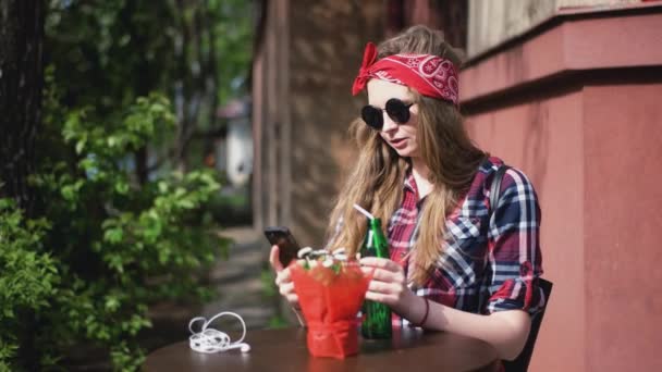 Urban nastoletnich Hipster dorywczo szczęśliwy dziewczyna w okularach słonecznych i barwna chustka uśmiech i napić się wody na tarasie w słoneczny dzień — Wideo stockowe