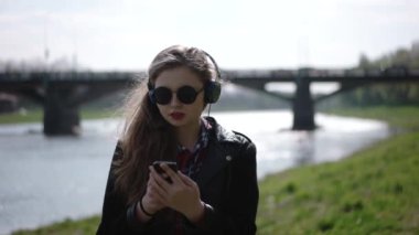 Oturma ve Nehri yakınında yeşil çimenlerin üzerinde rahat müzik dinlemek güneş gözlüklü genç Casual Kız