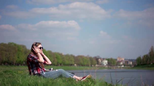 Casual tienermeisje in zonnebril luisteren naar de muziek zitten en ontspannen op een groen gras in de buurt van de rivier — Stockvideo