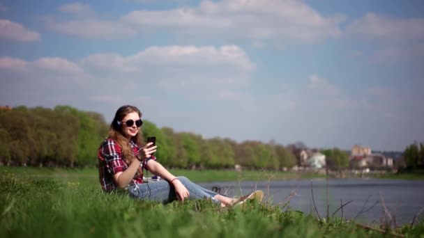 Adolescente chica casual en gafas de sol escuchando la música sentada y relajada en una hierba verde cerca del río — Vídeos de Stock