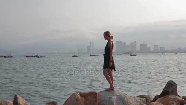 Νεαρή γυναίκα στέκεται στην παραλία στις πέτρες και βλέποντας πάνω στη θάλασσα — Αρχείο Βίντεο