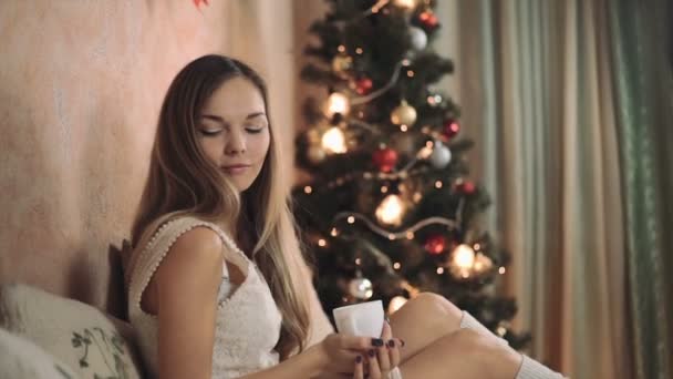 Mujer en suéter caliente en la cama con taza de café en las manos, árbol de Navidad nuevo año — Vídeo de stock