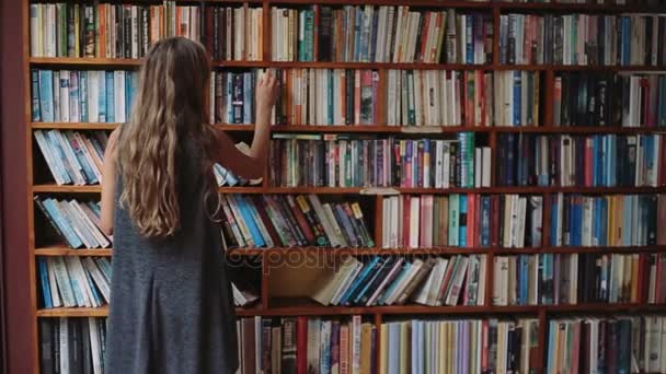 Beautiful young girl with book in hand walks along the bookshelves — Stock Video