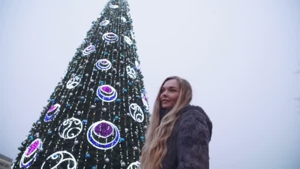 Donna attraente la notte di Natale sorride guardando la fotocamera di fronte agli alberi del parco decorate luci scintillanti — Video Stock