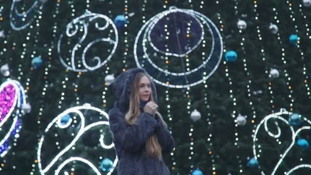 Mujer atractiva en la noche de Navidad sonríe mirando a la cámara delante de los árboles del parque decorado con luces brillantes — Vídeos de Stock