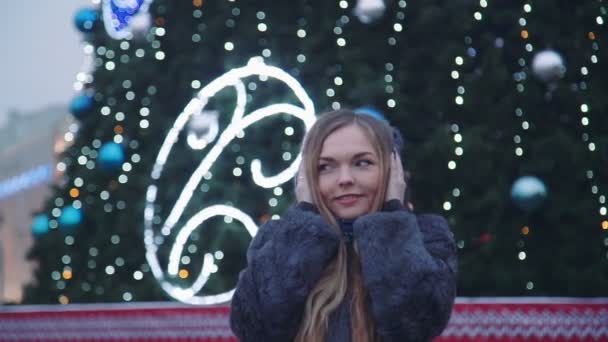 Mujer atractiva en la noche de Navidad sonríe mirando a la cámara delante de los árboles del parque decorado con luces brillantes — Vídeo de stock