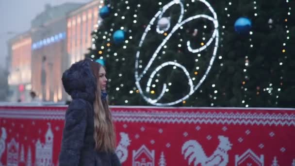 Привлекательная женщина проходит мимо рождественской елки и завернутая в улыбку перед садовыми деревьями, украшенными сверкающими огнями — стоковое видео