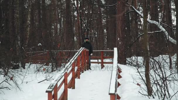 Молодой человек смотрит в камеру и играет со снегом в зимнем лесу. Он делает снежки и бросает их. Концепция зимних развлечений — стоковое видео