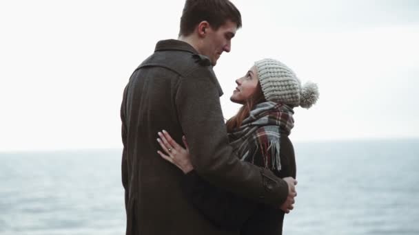 Pareja joven mirando al mar, abrazo en la playa, abrigo y sombrero, clima frío del Báltico o Islandia — Vídeos de Stock