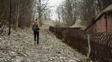 Sırt üstünde sırt çantası olan genç bir adam parkta bahar veya terk edilmiş bir Alman kale yanında sonbahar ağaçlar Sarı yaprakları tuğla duvarlar, eski bir demir çit yürür