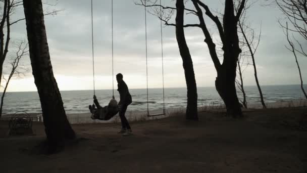 Young gelukkig lachend hipster fashion paar in liefde schommel op een schommel hangen aan een boom, met uitzicht op de zee, veel plezier buiten in de koude herfst lente weer poseren op de weg — Stockvideo