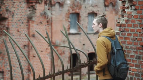 Sırt üstünde sırt çantası olan genç bir adam parkta bahar veya terk edilmiş bir Alman kale yanında sonbahar ağaçlar Sarı yaprakları tuğla duvarlar, eski bir demir çit yürür — Stok video
