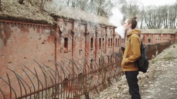 Egy fiatal ember, hátizsák vissza sétál a parkban a tavaszi vagy az őszi fák sárga levelek, mellett egy elhagyott német fort, tégla falak, egy régi vas kerítés — Stock videók