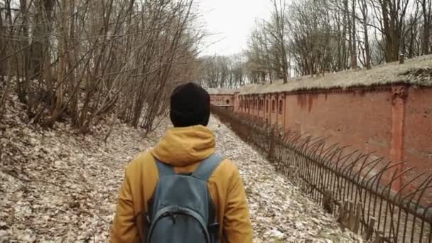 Un joven con mochila en la espalda camina por el parque en los árboles de primavera u otoño hojas amarillas, junto a un fuerte alemán abandonado, paredes de ladrillo, una antigua cerca de hierro — Vídeo de stock