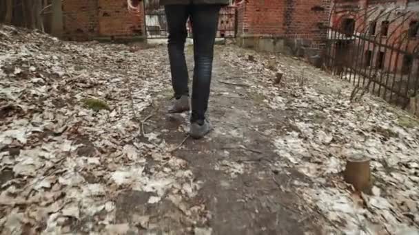 Gri paltolu bir sakallı genç bir adam parkta bahar veya terk edilmiş bir Alman kale yanında sonbahar ağaçlar Sarı yaprakları tuğla duvarlar, eski bir demir çit yürür — Stok video