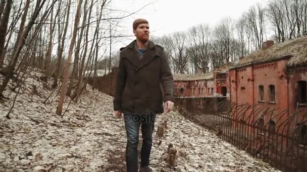 Egy fiatal férfi szakáll, egy szürke kabátot sétál a parkban a tavaszi vagy az őszi fák sárga levelek, mellett egy elhagyott német fort, tégla falak, egy régi vas kerítés — Stock videók
