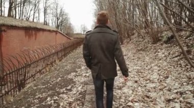 Gri paltolu bir sakallı genç bir adam parkta bahar veya terk edilmiş bir Alman kale yanında sonbahar ağaçlar Sarı yaprakları tuğla duvarlar, eski bir demir çit yürür