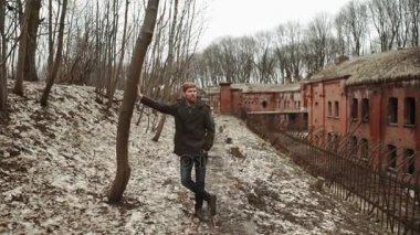 Gri paltolu bir sakallı genç bir adam parkta bahar veya terk edilmiş bir Alman kale yanında sonbahar ağaçlar Sarı yaprakları tuğla duvarlar, eski bir demir çit yürür