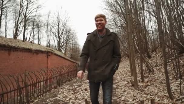 Un giovane con la barba, in un cappotto grigio cammina attraverso il parco in primavera o autunno alberi foglie gialle, accanto a un forte tedesco abbandonato, muri di mattoni, un vecchio recinto di ferro — Video Stock