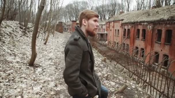 Un joven con barba, con un abrigo gris camina por el parque en los árboles de primavera u otoño hojas amarillas, junto a un fuerte alemán abandonado, paredes de ladrillo, una antigua cerca de hierro — Vídeos de Stock