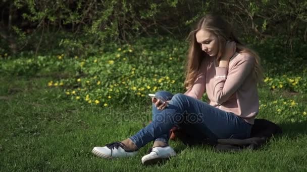 Schöne junge Frau in Jeans sitzt glücklich im sattgrünen Gras in einem Sommerpark unter sonnenbeschienenen Bäumen — Stockvideo
