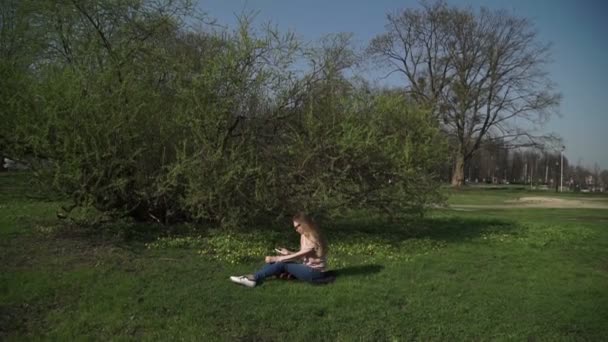 Hermosa joven en jeans sentada felizmente en la exuberante hierba verde en un parque de verano bajo árboles soleados — Vídeos de Stock