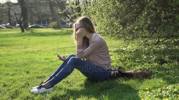 Mulher bonita em jeans sentada feliz em exuberante grama verde em um parque de verão sob árvores iluminadas pelo sol — Vídeo de Stock