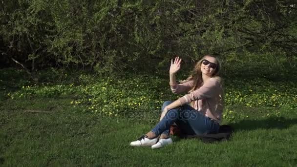 Beautiful young woman in jeans sitting happily in lush green grass in a summer park under sunlit trees — Stock Video