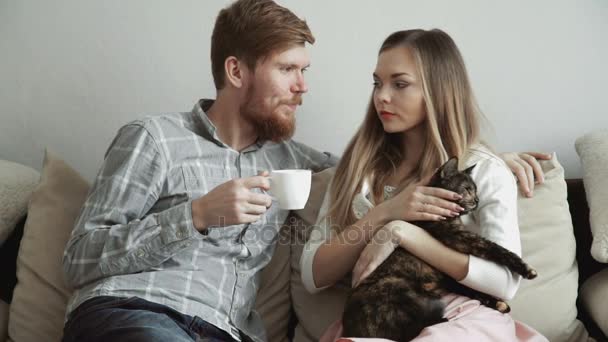 Pareja está sentado en el sofá del sofá en casa acariciando a un gato, beber té de un juego de té blanco y ver la televisión . — Vídeos de Stock