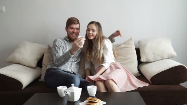 Paar sitzt zu Hause auf dem Sofa und schaut ins Smartphone und lächelt, trinkt Tee aus einem weißen Tee-Set und schaut fern. — Stockvideo