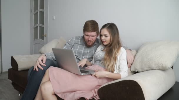 Pareja está sentada en el sofá del sofá en casa Mira la pantalla del ordenador portátil y sonríe — Vídeos de Stock