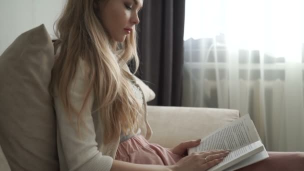 Schöne junge Frau sitzt auf Sofa und liest Buch und lächelt — Stockvideo