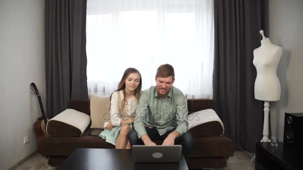 Couple is sitting on the couch sofa at home Look at the laptop screen and smile — Stock Video