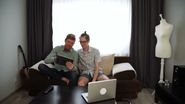 Deux jeunes gars frères travaillent de la maison assis sur un canapé et de travailler sur sa tablette, sur un ordinateur portable de table . — Video