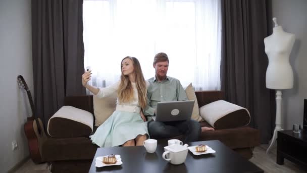Ζευγάρι κάθεται στον καναπέ καναπέ στο σπίτι κάνει selfie φωτογραφία στο smartphone ματιά στην οθόνη φορητού υπολογιστή και χαμόγελο — Αρχείο Βίντεο