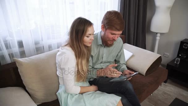 Paar sitzt zu Hause auf dem Sofa und schaut in das Tablet und lächelt aus nächster Nähe — Stockvideo