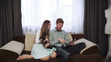 Çift evde kanepe kanepede göz oturuyor tablet ve gülümseme, içecek çay beyaz çay seti ve tv izleme içine.