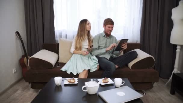 Pár ül otthon a kanapén kanapén megjelenés a tabletta és a mosoly, ital tea, fehér tea-set és a tévénézés. — Stock videók