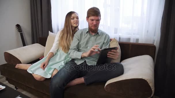 Pár sedí na gauči pohovce doma podívat do tabletu a úsměv, pití čaje bílý čaj set a dívat se na televizi. — Stock video