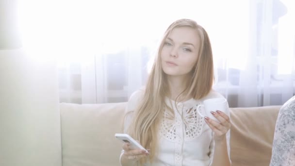 Hem, teknik och internet koncept - leende kvinna som sitter i soffan med smartphone hemma. Dricka te från en vit teservis. Solen bländning, mot solen ljus som lyser — Stockvideo