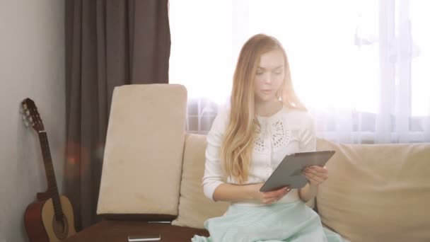 Maison, technologie et concept internet - femme souriante assise sur le canapé avec tablette à la maison. Buvez le thé d'un ensemble de thé blanc. Éblouissement du soleil, Contre le soleil lumières vives — Video