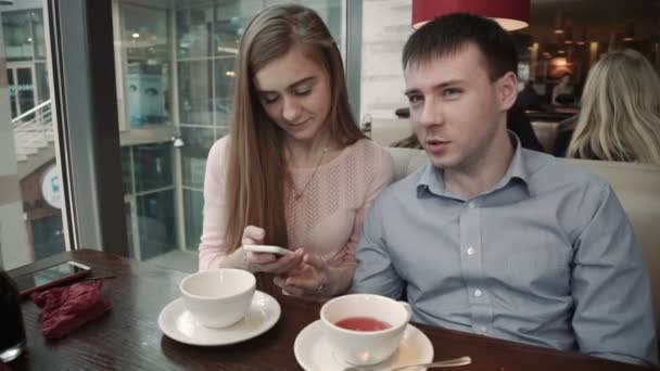 Atractiva pareja joven utilizando el teléfono inteligente en la cafetería Mira la pantalla sonriendo — Vídeo de stock