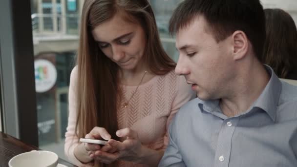 Atractiva pareja joven utilizando el teléfono inteligente en la cafetería Mira la pantalla sonriendo — Vídeos de Stock