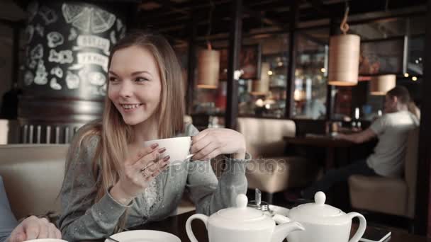 Молодая женщина ждет кого-то в кафе. Она пьет чай и разговаривает с друзьями. Улыбаясь и глядя в камеру — стоковое видео