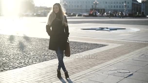 Молода красива жінка в сірому пальто з довгим волоссям, посміхаючись і позує на модель моди камери в центрі міста на заході сонця, тепле світло і відблиски на лінзі — стокове відео
