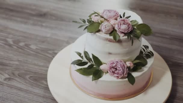 Een twee-voudige wedding cake, versierd met takjes groen en verse bloemen met rozen en pioenrozen, staat op een houten tafel — Stockvideo