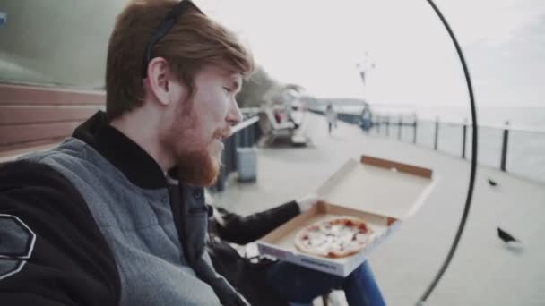 Pár pizzát eszik a tengerparton, hideg időjárás, együtt. Fiatal férfi kabát és a nő egy kabátot, egy padon a mólón, néz a tengerre — Stock videók