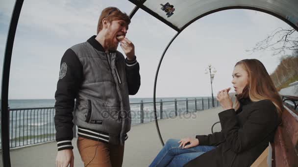 Paar pizza op de kust, koud weer, samen eten. Jonge man in de jas en de vrouw in een vacht, op een bankje bij de pier kijken naar de zee — Stockvideo